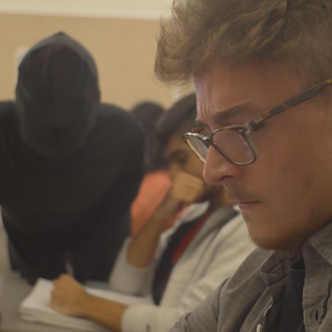 A man with glasses in a classroom, staring off frame as a humanoid, shadow figure looms over him.